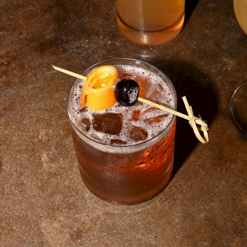 A fancy drink in a rounded glass with a cherry garnish and a lemon slice, served on a bar counter with a straw.