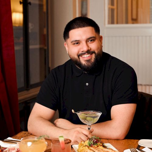 A man sits at a dining table with plates of food, looking at the camera in a cozy, warmly lit setting.
