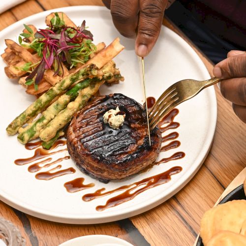 A chocolate donut with a drizzle of sauce, a small pastry, and a few colorful bites on a wooden board, with hands in the background.