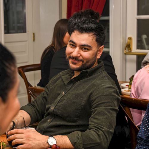 A man smiles while playing a card or board game with others at a table; warm, cozy room with red curtains in the background.