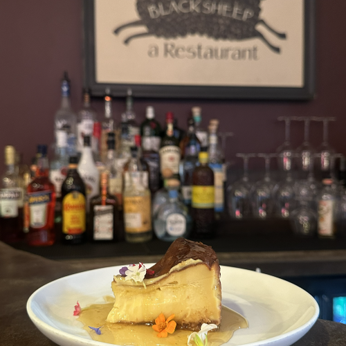 A dessert plate with a slice of cake and a scoop of ice cream, set against a bar with bottles and a vintage black-and-white mural in the background.