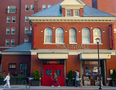 A brick hotel with a blue roof and arched windows; sign reads &ldquo;The Kendall Hotel&rdquo; with people walking by and a red door.