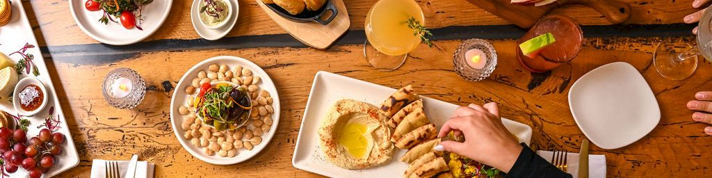 A bustling shared meal: hands reach for skewers, chips and bread, a platter of cheeses, olives, and nuts, plus drinks and dessert on a rustic wooden table.