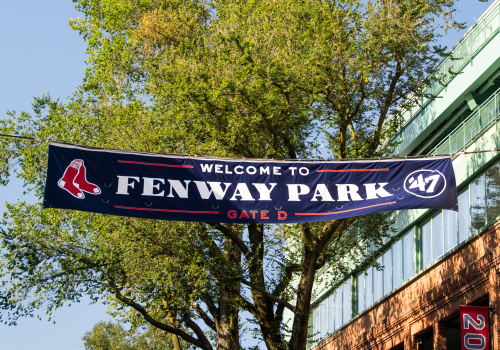 A banner reads &ldquo;Welcome to Fenway Park&rdquo; with Gate D, hanging above a street near a brick building and green trees on a sunny day.