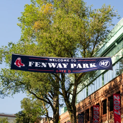 A banner reads &ldquo;Welcome to Fenway Park&rdquo; with Gate D, hanging above a street near a brick building and green trees on a sunny day.