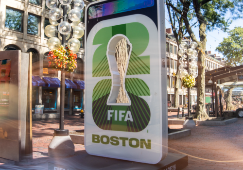 A tall, glowing FIFA World Cup display for Boston, featuring a trophy image, surrounding trees, and urban storefronts in a sunny plaza.