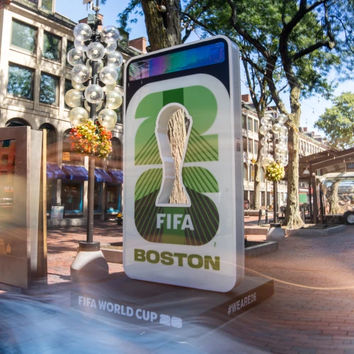 A tall, glowing FIFA World Cup display for Boston, featuring a trophy image, surrounding trees, and urban storefronts in a sunny plaza.