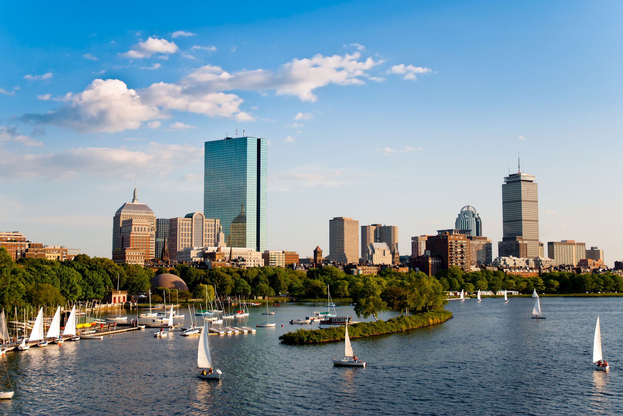 A city skyline along a river with sailboats, green trees lining the shore, and a bright blue sky with scattered clouds.