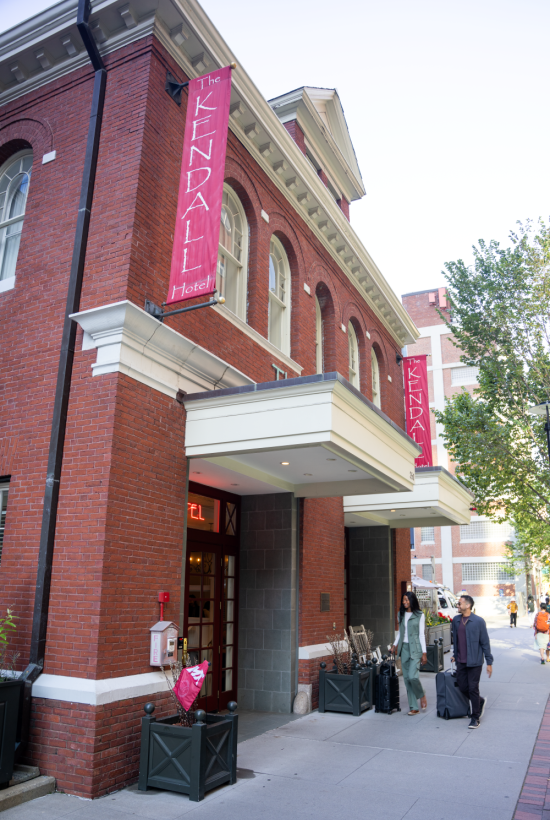 A brick theater or venue on a corner with magenta banners reading &ldquo;KINDAI&rdquo; or similar, people walking outside the entrance.