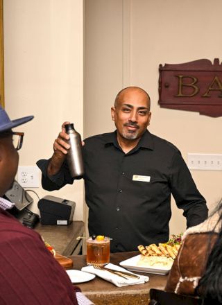 A man in a suit sits at a desk with two people facing him; a woman in a dark dress on the right, a person in a hat on the left, a small office setting.