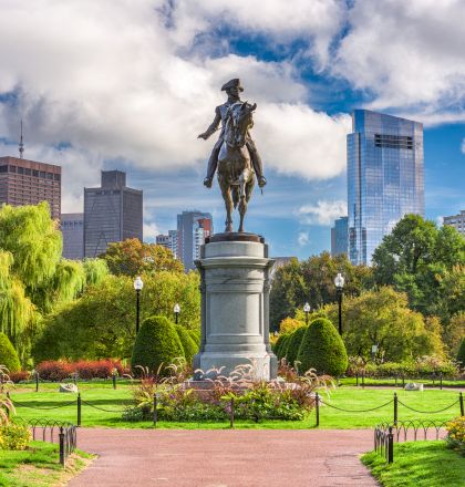 A statue in a park with a fountain and manicured gardens, blue sky, and green trees in the background.