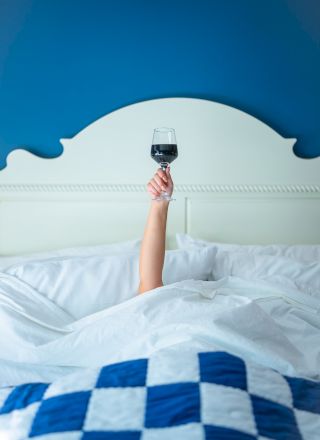 A person lies in bed under a white comforter, arm raised above the blanket holding a glass of red wine, with a blue painted wall behind.