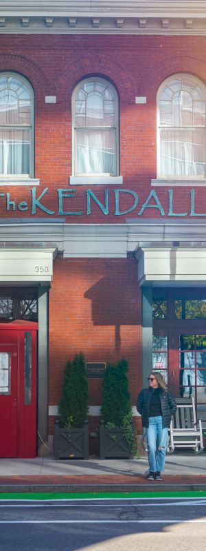 A brick storefront with a blue &ldquo;The Kendal&rdquo; sign, red door on the left, blue door on the right, a person walking past, and street lamps.