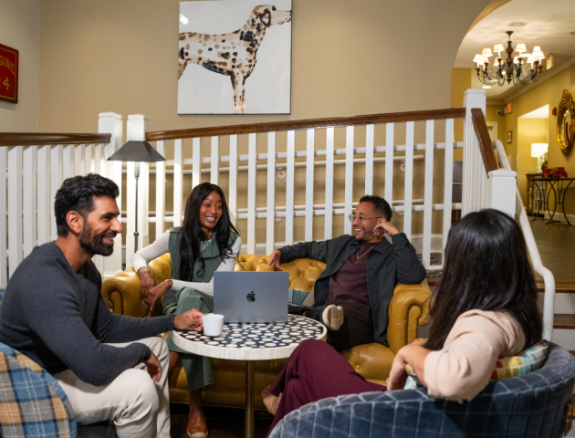 Two friends sit at a cozy cafe table chatting and laughing, with warm lights, plants, and shelves in the background.