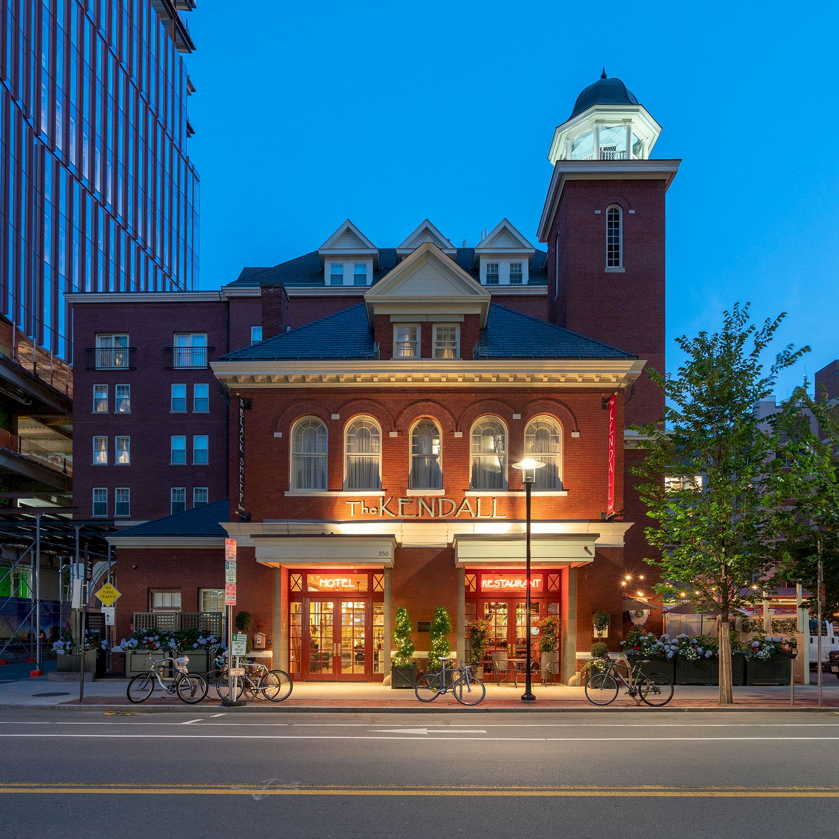 A historic red-brick building with a clock tower, storefronts at street level, bicycles parked out front, and modern buildings flanking it on a sunny day.