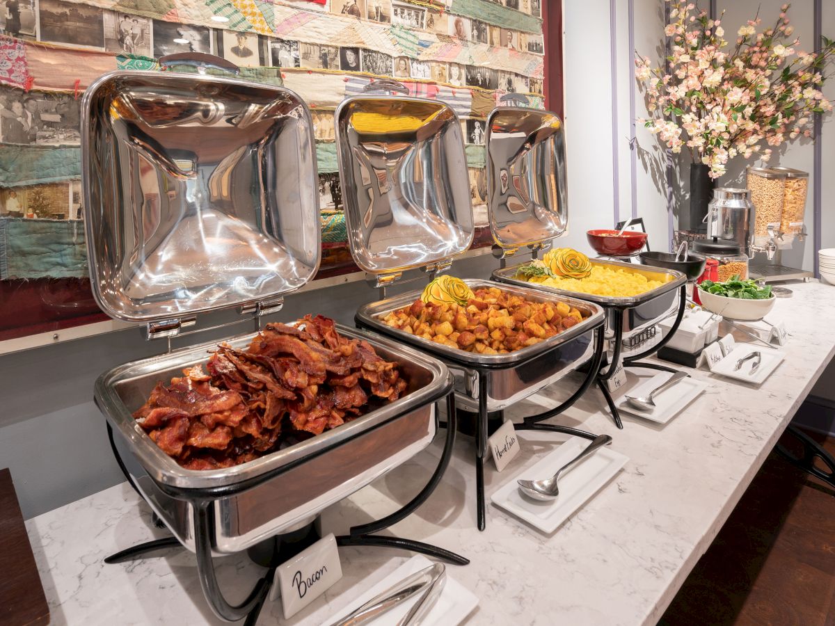 A buffet with chafing dishes of cooked meats and sides on a marble counter, bowls, plates, and decorative flowers in the background.