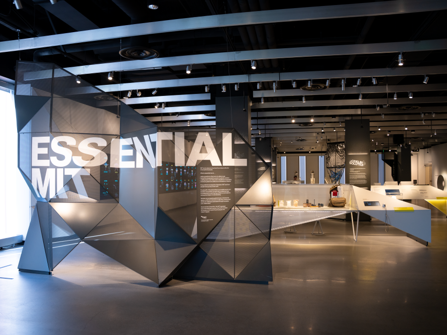 A modern showroom with geometric gray walls reading &ldquo;ESSENTIAL&rdquo; near the entrance, sleek white tables, bright lighting, and dark ceiling beams.