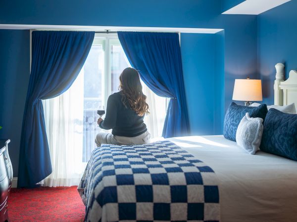 A woman sits by a bright window with blue curtains in a vivid, blue-themed hotel room, checkerboard bedspread, and cozy pillows, watching the view.