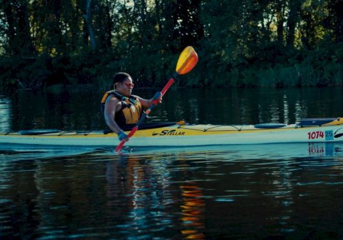 A person kayaking on calm water, wearing a life jacket, using a double-blade paddle; trees line the background.