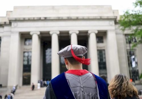 A graduate in a cap and gown stands facing a grand columned building, likely at a commencement ceremony, with onlookers nearby.
