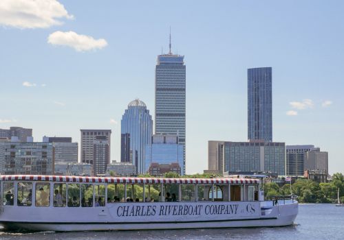 A river cruise boat labeled &ldquo;Charles Riverboat Company&rdquo; sails by a city skyline with tall modern buildings in the background.