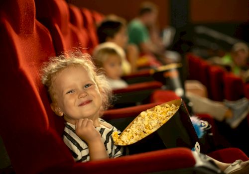 A smiling little girl sits in a red theater seat, clasping her chin, with a bucket of popcorn, others blurred in the background.