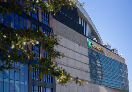 A large modern building with glass windows and a TD Garden sign on the side, partially shaded by trees against a clear blue sky.