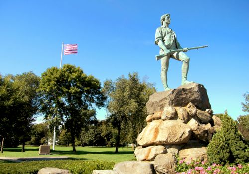 A statue of a soldier with a rifle stands on a rocky pedestal in a park, American flag waving, trees and flowers around.