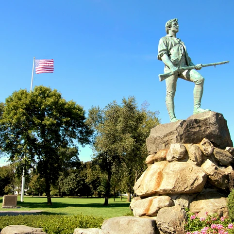 A statue of a soldier with a rifle stands on a rocky pedestal in a park, American flag waving, trees and flowers around.