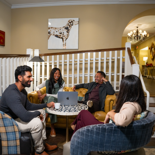 A group of four casually dressed friends sit around a coffee table in a cozy lounge, chatting and laughing inside a warmly lit, stylish living room.