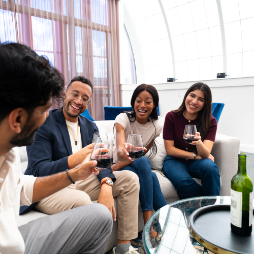 Four friends share drinks and laughter in a bright lounge, clinking glasses around a glass coffee table with a bottle on it.