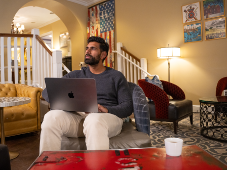 A man sits with a laptop in a cozy, art-filled living room, neutral tones, a red coffee table, and soft lighting in the background.