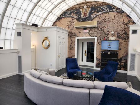 Luxurious modern living room with a curved white sofa, blue chairs, a grand map mural, glass ceiling, and a cozy media setup.