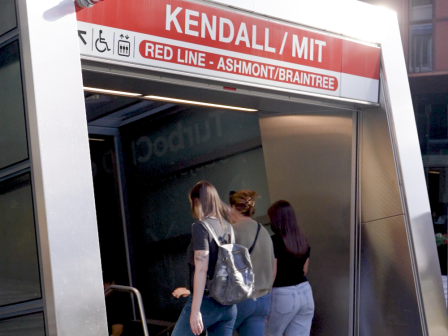 People ride the red line subway into a modern station entrance labeled &ldquo;KENDALL / MIT,&rdquo; with a red banner and glass buildings in the background.