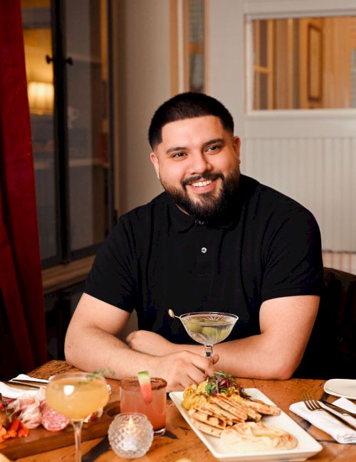 A smiling man with a beard sits at a dining table with drinks, appetizers, and a plate of food in a cozy restaurant setting.