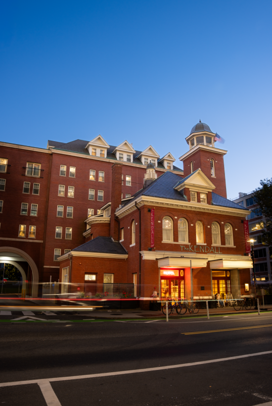 A brick hotel with a tower and lit storefront, curved entrance, and a blue sky at dusk, bustling street below.