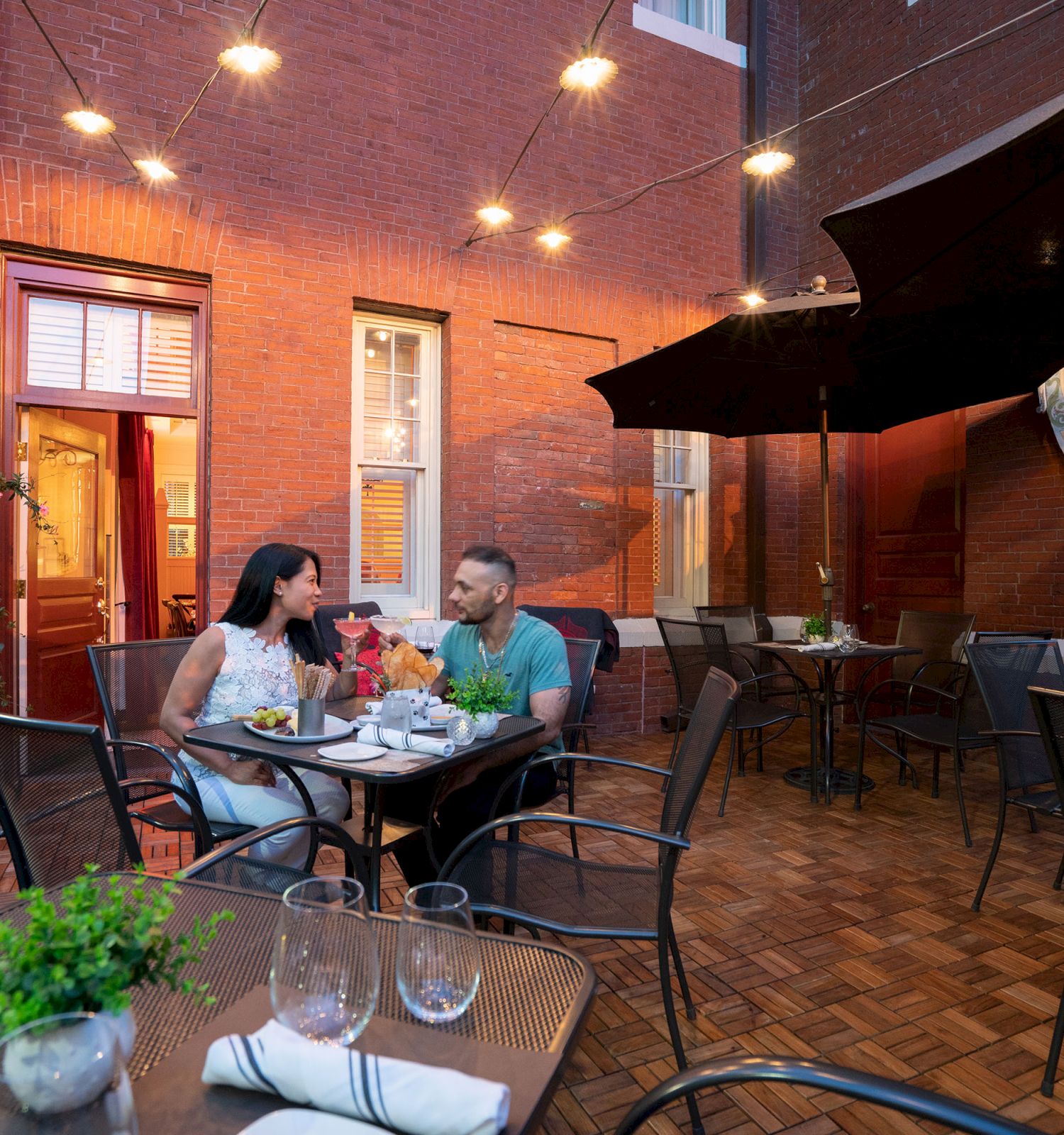 Two people dine al fresco at a cozy brick courtyard restaurant, candlelit tables, potted plants, and string lights overhead.