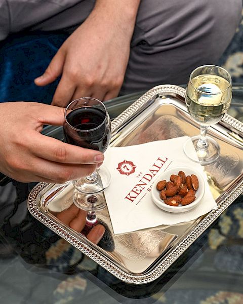 Two people sharing a drink tray: a hand holding a glass of red wine, another hand nearby, with a small plate of almonds and a white card on a silver tray.