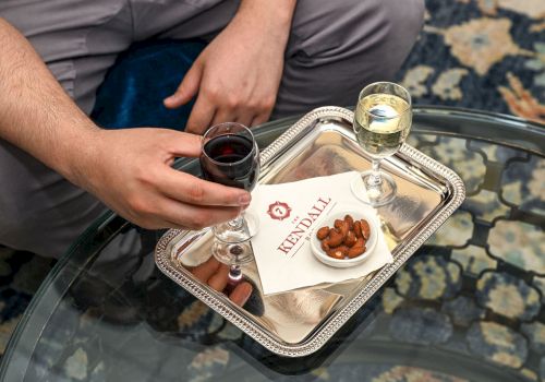 Two people sharing a drink tray: a hand holding a glass of red wine, another hand nearby, with a small plate of almonds and a white card on a silver tray.
