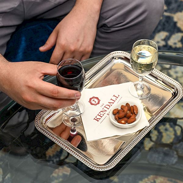 Two people sharing a drink tray: a hand holding a glass of red wine, another hand nearby, with a small plate of almonds and a white card on a silver tray.
