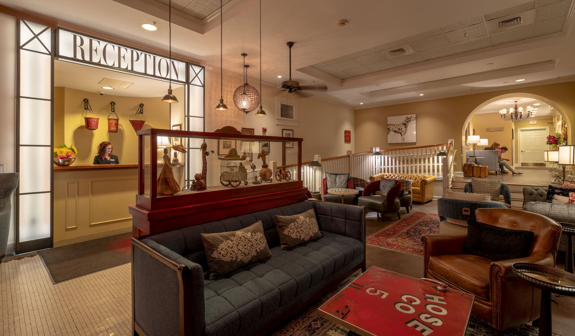 Hotel lobby with a reception desk, &ldquo;RECEPTION&rdquo; sign, lounge seating, chandeliers, and a glass display case.