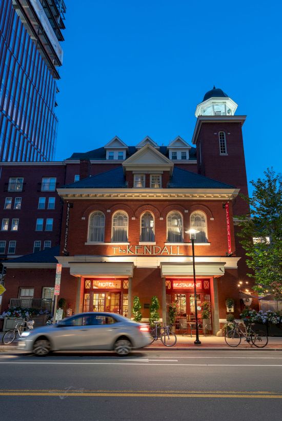A brick building with a tower is illuminated at night, with cars passing by on the street in front of it.