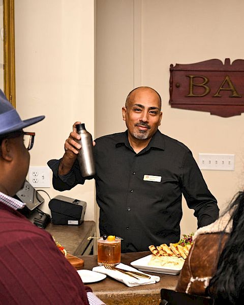 A bartender in a dark shirt pours a drink with a shaker while customers sit at a bar with snacks.