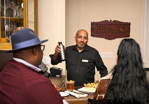 A bartender in a dark shirt pours a drink with a shaker while customers sit at a bar with snacks.