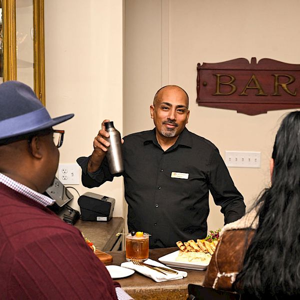 A bartender in a dark shirt pours a drink with a shaker while customers sit at a bar with snacks.