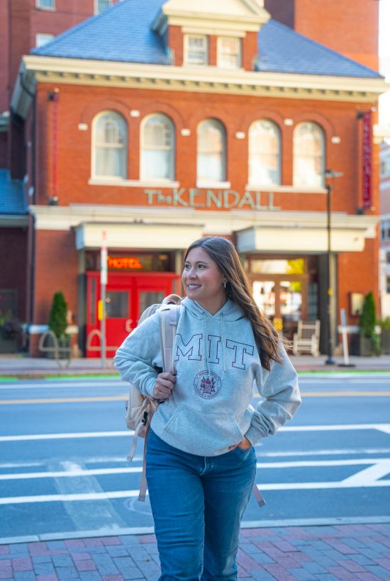A person in a casual outfit stands on the street near a brick building with a sign reading "KENDALL" in the background.