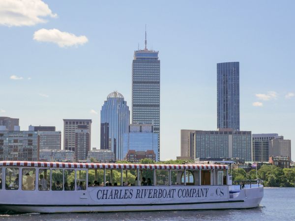 A Charles Riverboat Company tour boat cruises past Boston&rsquo;s skyline, with tall skyscrapers behind it.