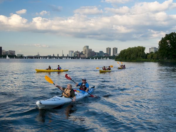 People paddle whitewater-style kayaks on a wide river, with city buildings and sailing boats in the distance under clouds.