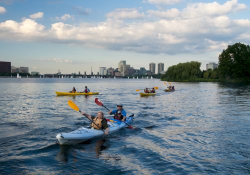 People kayak on a river under a cloudy sky, with a city skyline and sailboats in the distance.