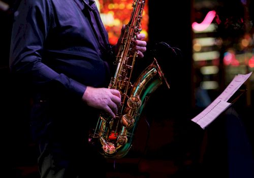 A musician in a dark suit holds a shiny saxophone while reading sheet music under colorful stage lights.